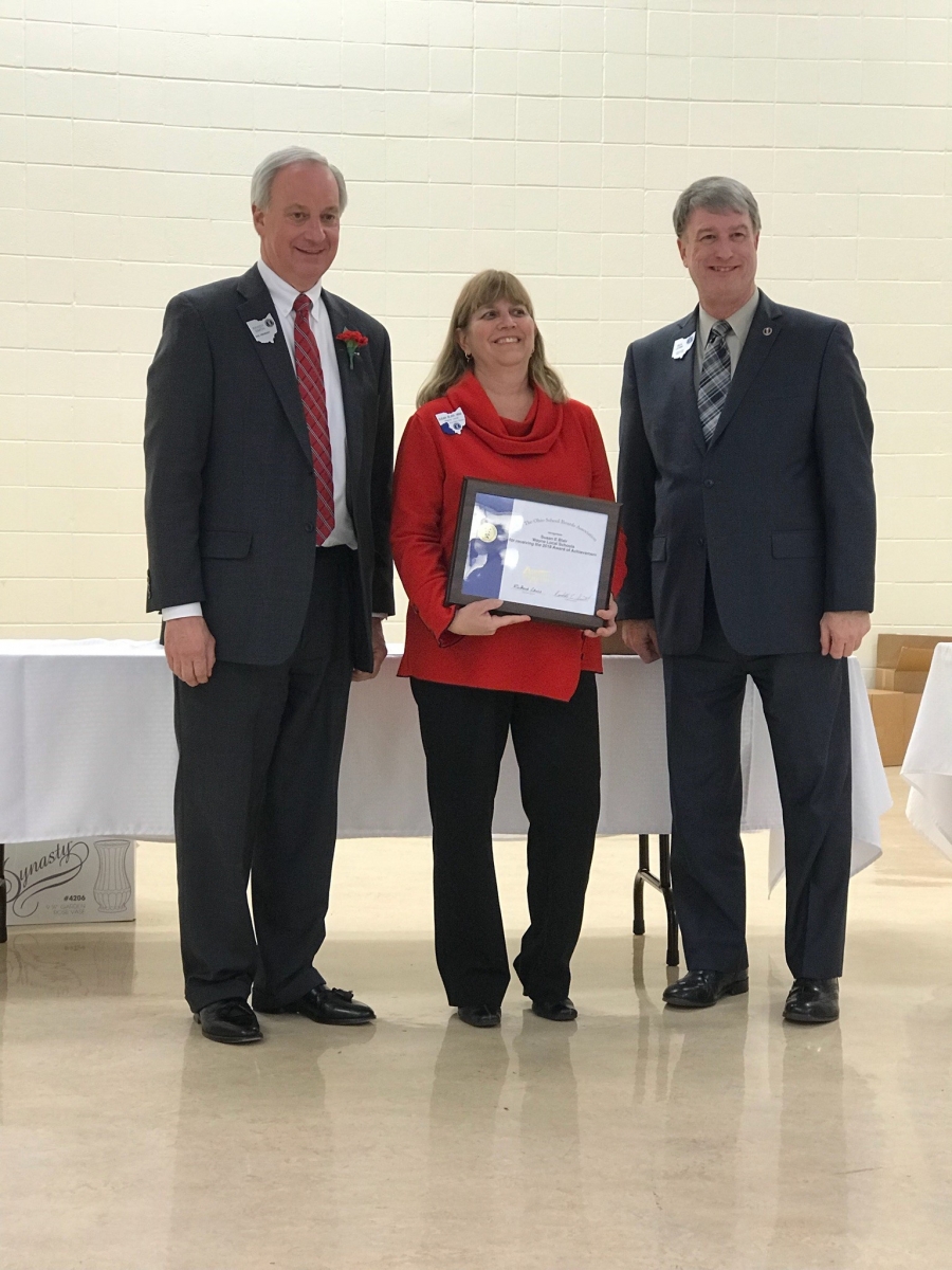 two men wearing suits with a woman in a red sweater in between them holding a plaque